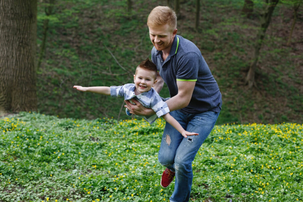 Pai brincando de aviãozinho com o filho em um parque florido, transmitindo segurança na Psicokids ABC.