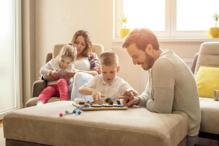 Menino brincando com jogo educativo enquanto o pai observa e a mãe cuida da irmã ao fundo na Psicokids ABC.