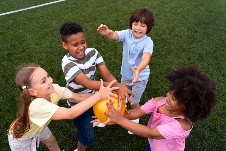 Quatro crianças diversas sorridentes brincando juntas com uma bola amarela em um gramado na Psicokids ABC.
