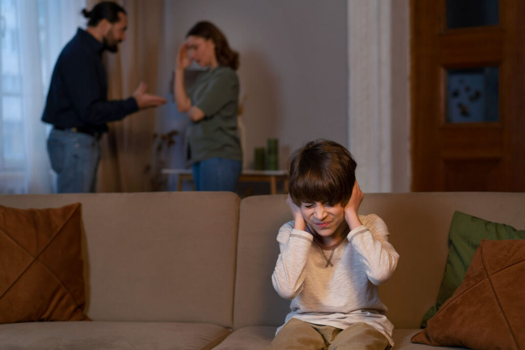 Menino tapando os ouvidos no sofá enquanto um casal discute ao fundo. A Clínica Psicokids ABC auxilia no impacto da separação dos pais.